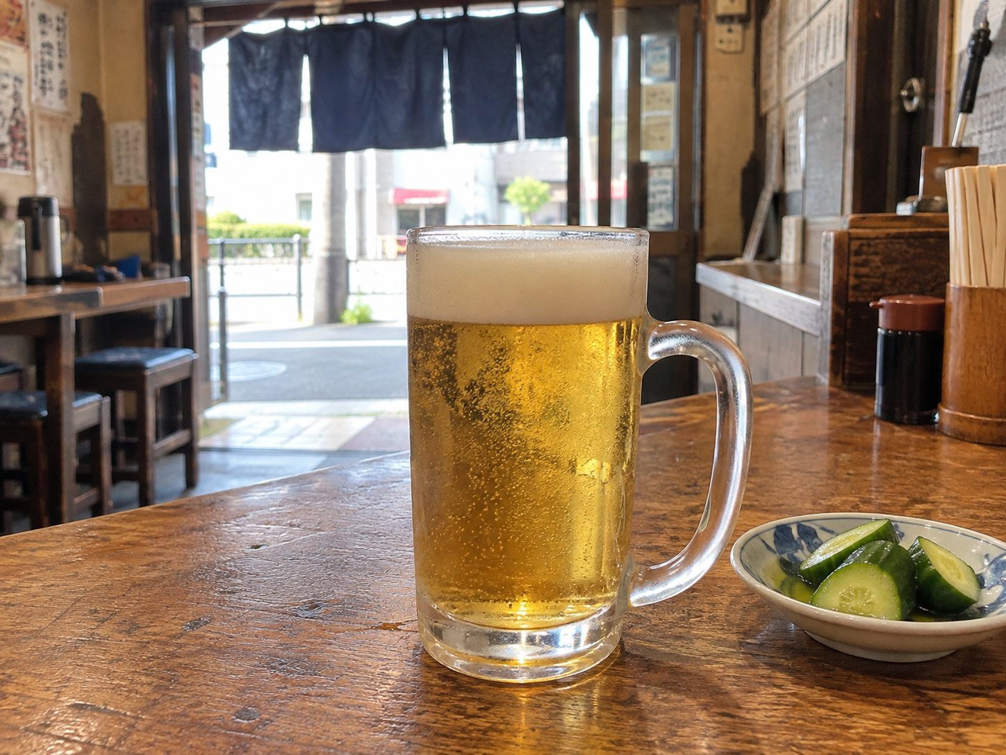 A beer mug on an izakaya counter in daylight, with the open door and noren curtain behind