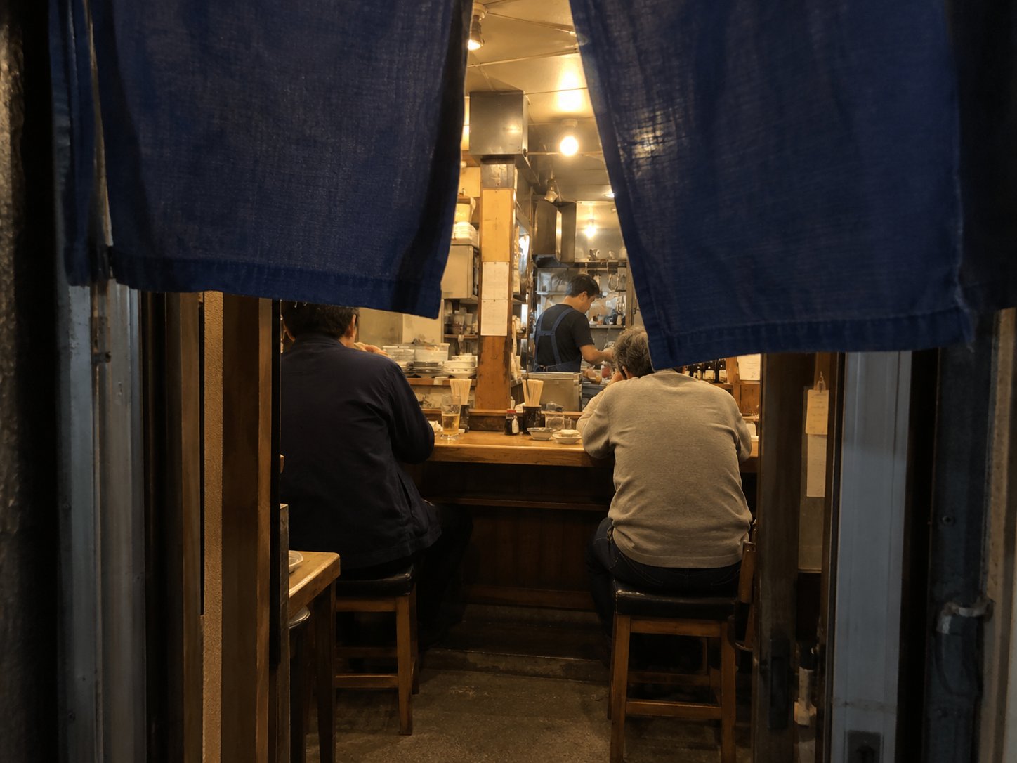 Looking into a small izakaya through a parted noren curtain, with customers at the counter and a cook in the back