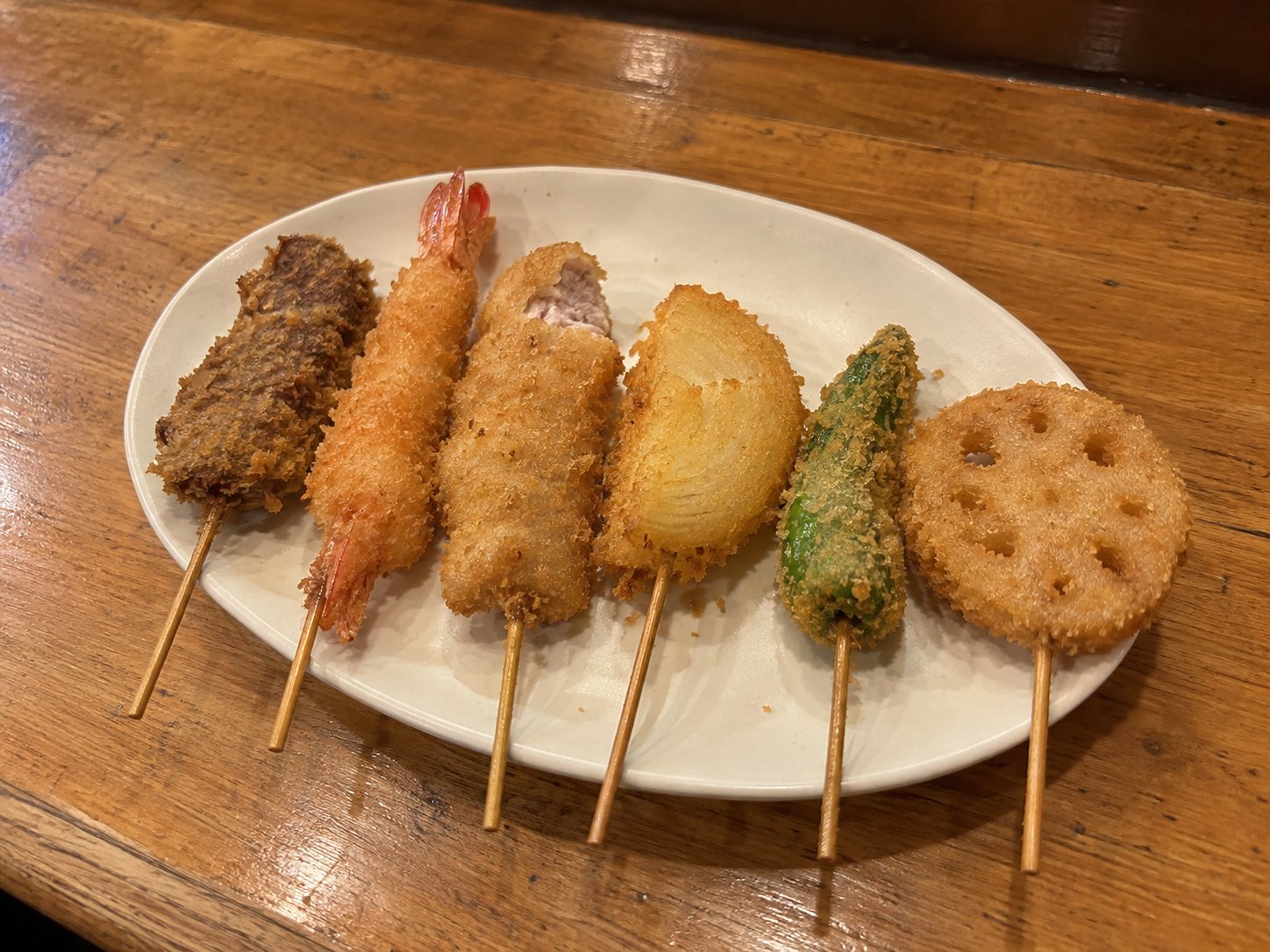 Six kushikatsu skewers on an oval plate: meat, prawn, onion, green pepper and lotus root, one with a bite taken out