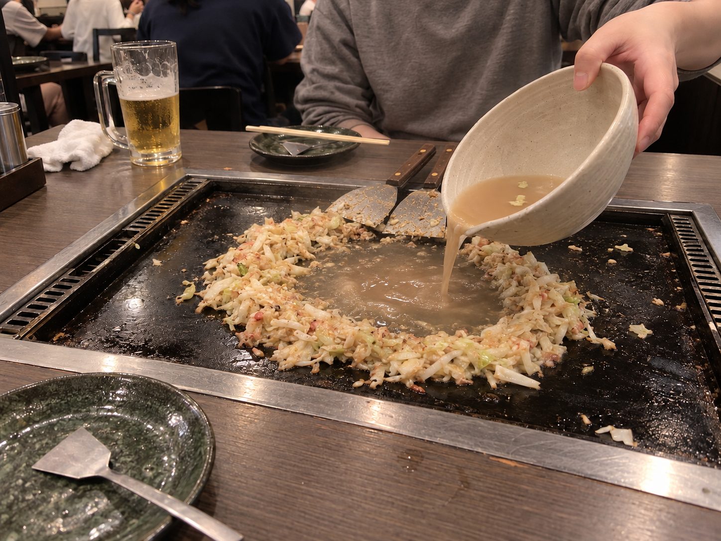 Batter being poured from a bowl into the middle of the ring of cabbage on the hot plate