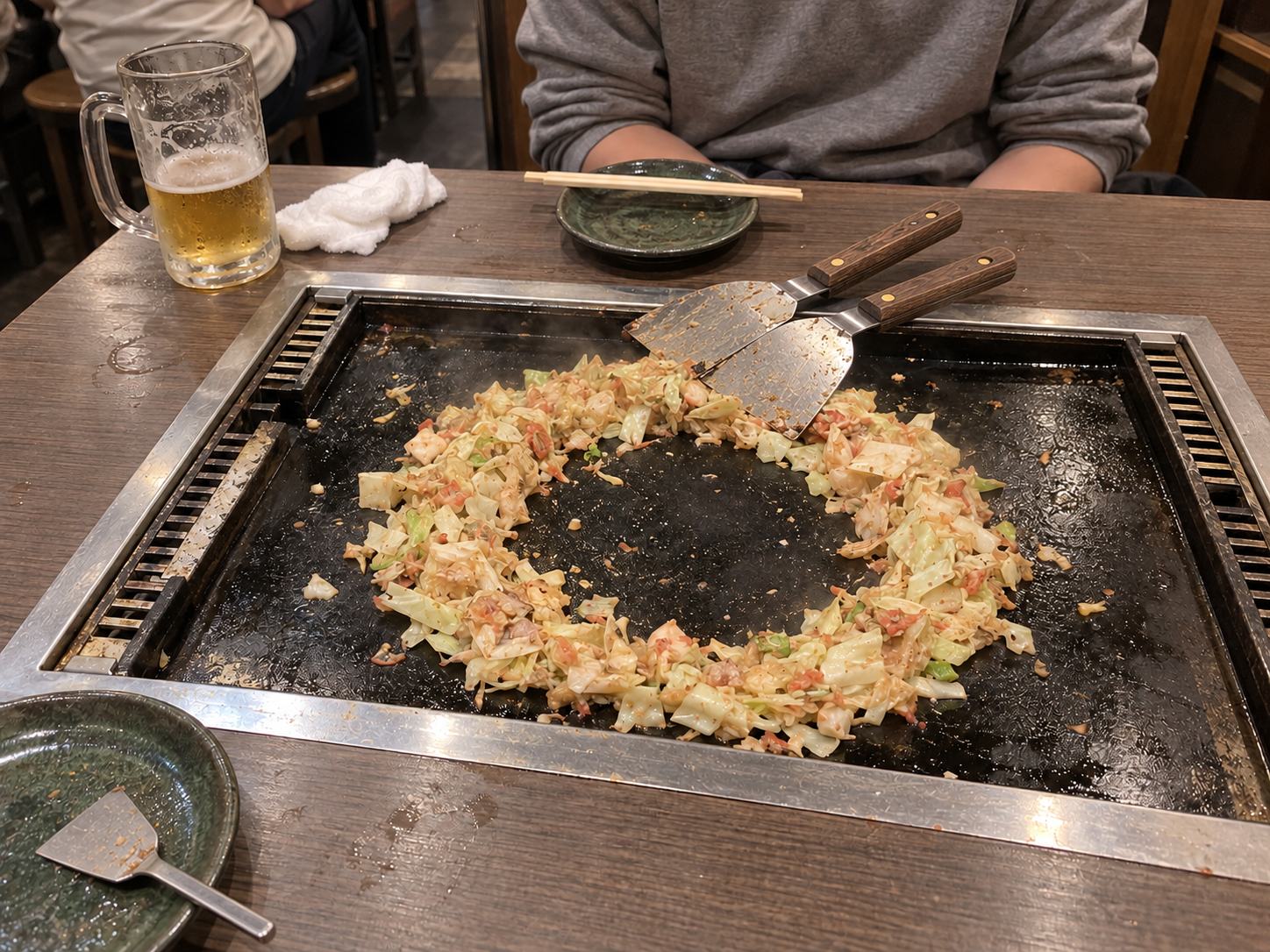 Fried cabbage and toppings shaped into a ring on the hot plate, with two large spatulas