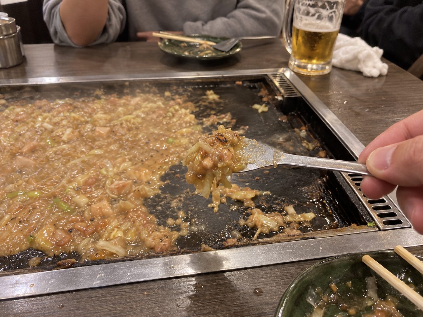 A hand holding a small spatula with a bite of monjayaki on it, above the hot plate