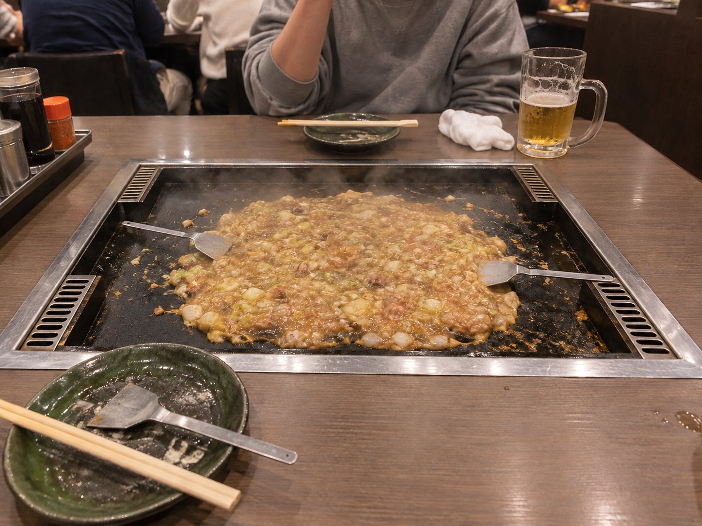 A table with a hot plate holding a thin layer of monjayaki with two small spatulas, next to a mug of beer