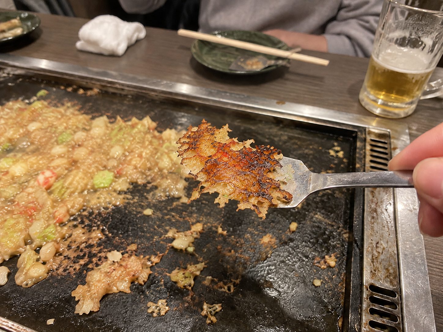 A small spatula holding a lacy, browned crisp of monjayaki above the hot plate