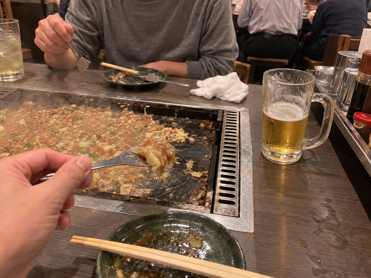 A hand lifting a bite of monjayaki with a small spatula, next to a mug of beer