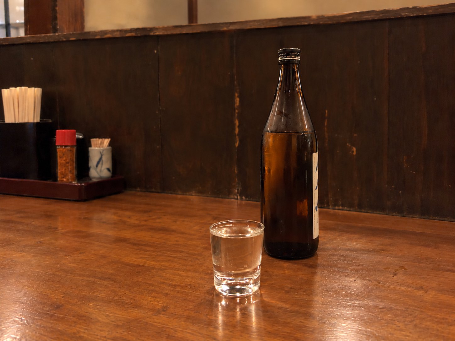 A small glass of sake on a wooden counter next to a brown bottle