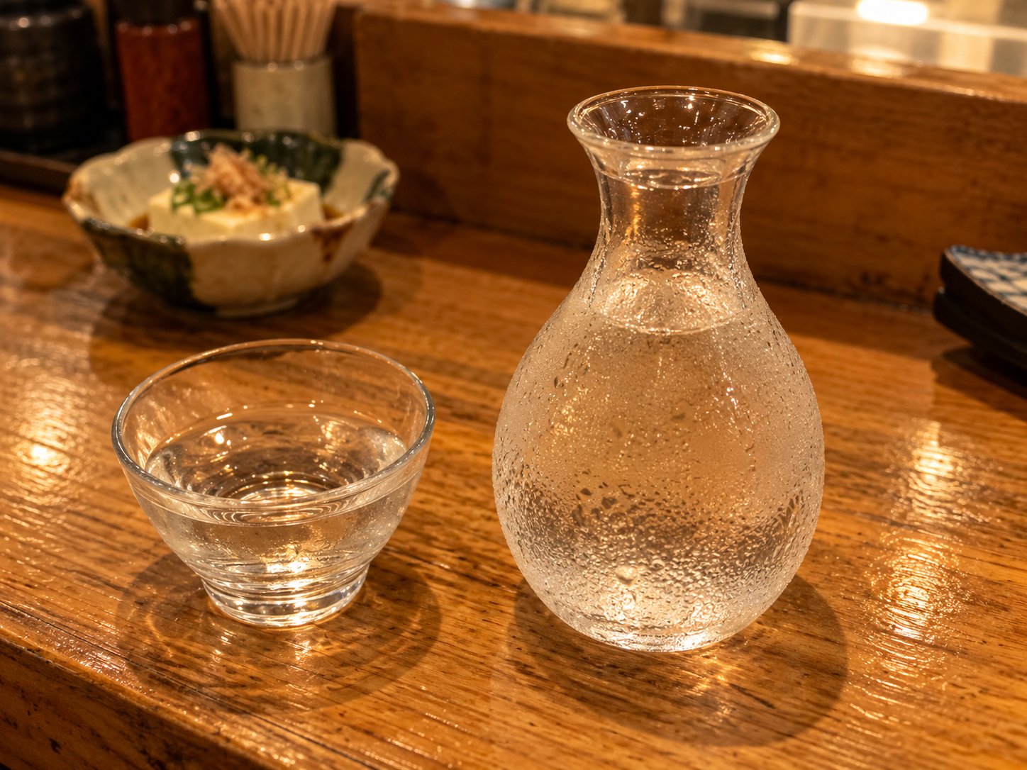 A glass sake flask covered in drops of condensation, next to a small glass cup