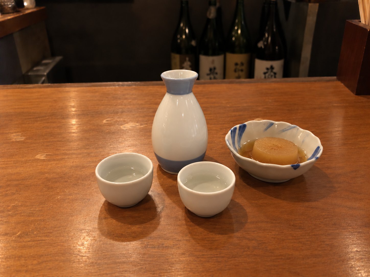 A white sake flask with a blue band and two small cups on a wooden counter, next to a bowl of simmered daikon